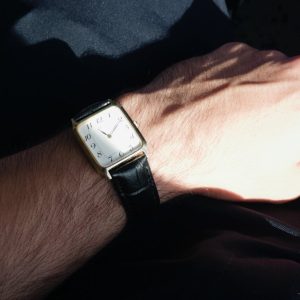 Stylish square wristwatch with leather band on wrist, captured in sunlight.